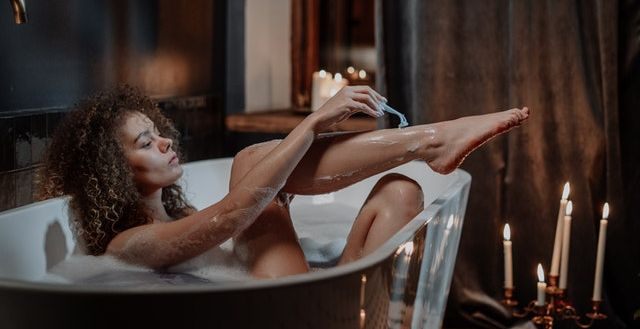 a lady shaving her legs lying on a bathtub