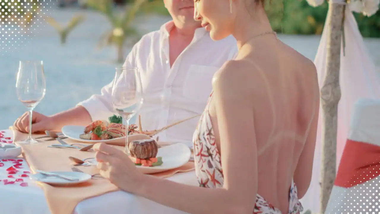 This image shows a couple sitting at an outdoor table, There are two wine glasses on the table in front of them and some food. there is a woman with tan lines on her back